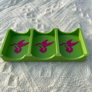 Puede incluir: Una bandeja verde lima de tres secciones con un dise&ntilde;o de colibr&iacute; rosa en cada secci&oacute;n. La bandeja est&aacute; hecha de un material s&oacute;lido y descansa sobre una superficie blanca texturizada. El dise&ntilde;o del colibr&iacute; es una silueta estilizada.