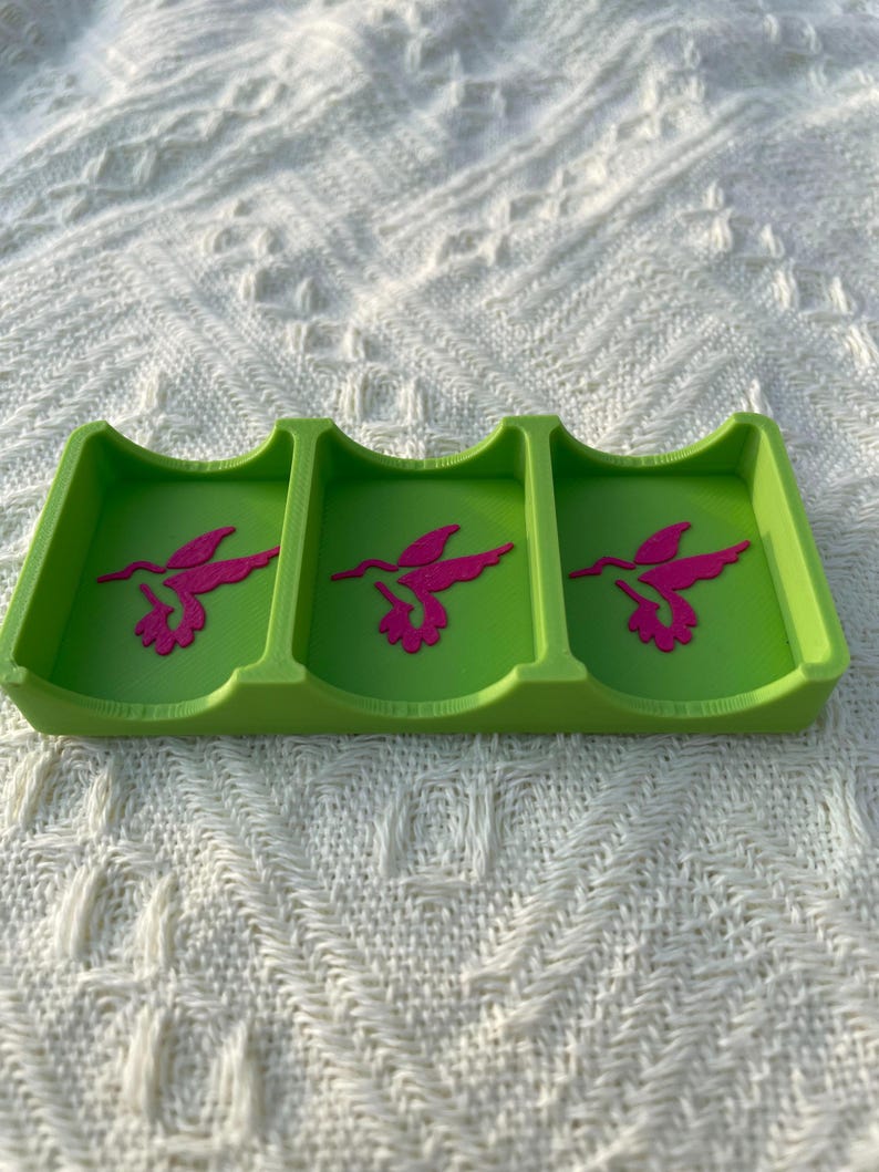 Puede incluir: Una bandeja verde lima de tres secciones con un dise&ntilde;o de colibr&iacute; rosa en cada secci&oacute;n. La bandeja est&aacute; hecha de un material s&oacute;lido y descansa sobre una superficie blanca texturizada. El dise&ntilde;o del colibr&iacute; es una silueta estilizada.