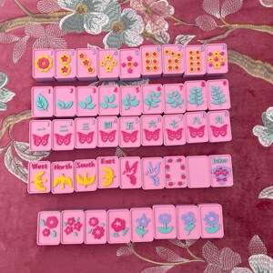 May include: A set of pink Mahjong tiles with various symbols and numbers. The tiles feature floral, leaf, and butterfly designs, along with the words "West", "North", "South", and "East". A "Joker" tile is also present.