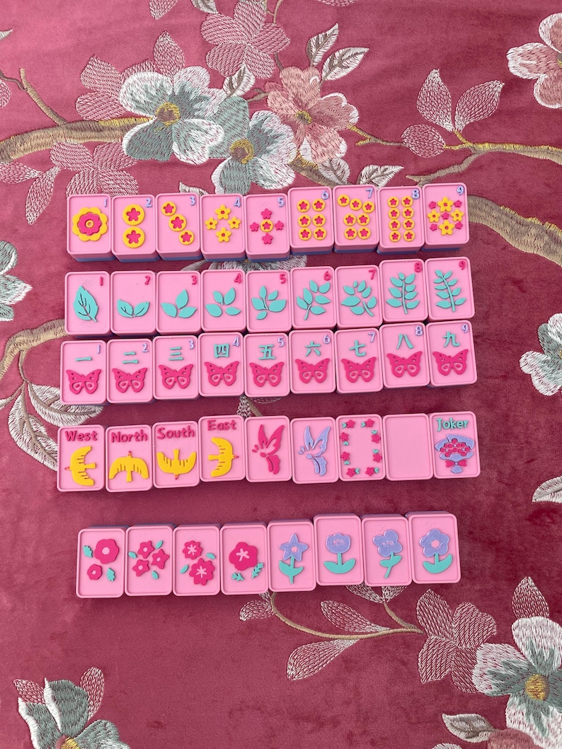 May include: A set of pink Mahjong tiles with various symbols and numbers. The tiles feature floral, leaf, and butterfly designs, along with the words "West", "North", "South", and "East". A "Joker" tile is also present.