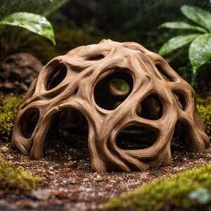 May include: A brown, porous, cave-like structure with multiple openings, resembling a natural formation. The object is set amidst green moss and foliage, creating a miniature landscape. The structure's color is a warm, earthy tone.