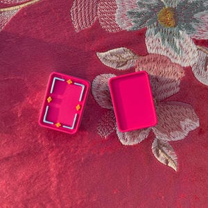 Puede incluir: Dos recipientes rectangulares de pl&aacute;stico rosa intenso. Un recipiente est&aacute; abierto, revelando un interior texturizado con un marco rectangular azul claro y cuatro peque&ntilde;os detalles en forma de estrella amarilla. El segundo es una tapa lisa.