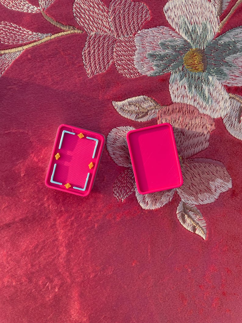 Puede incluir: Dos recipientes rectangulares de pl&aacute;stico rosa intenso. Un recipiente est&aacute; abierto, revelando un interior texturizado con un marco rectangular azul claro y cuatro peque&ntilde;os detalles en forma de estrella amarilla. El segundo es una tapa lisa.