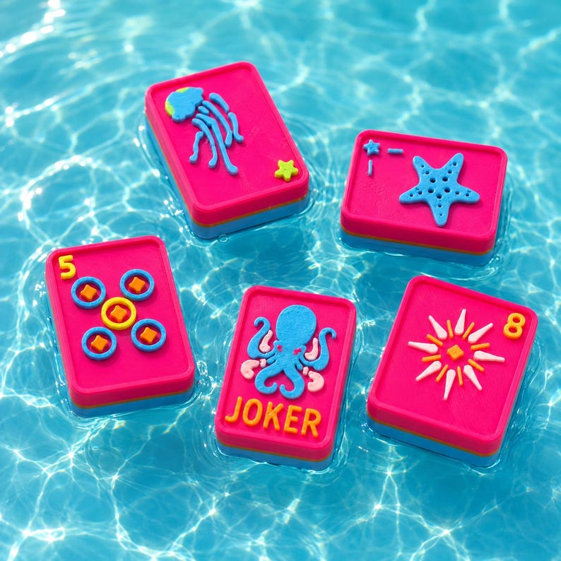 Puede incluir: Cinco fichas de juego rectangulares con superficies de color rosa brillante y bases azules. Las fichas presentan iconos acu&aacute;ticos coloridos en relieve, incluyendo una medusa, una estrella de mar, un pulpo con el texto JOKER, un n&uacute;mero 5 y un 8.
