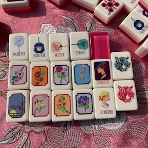 May include: A collection of vintage Mahjong tiles. The tiles are white with pink accents and feature various symbols, numbers, and characters. Some tiles display floral designs, while others have directional markers like "North" and "South". "Joker" tiles are also visible.