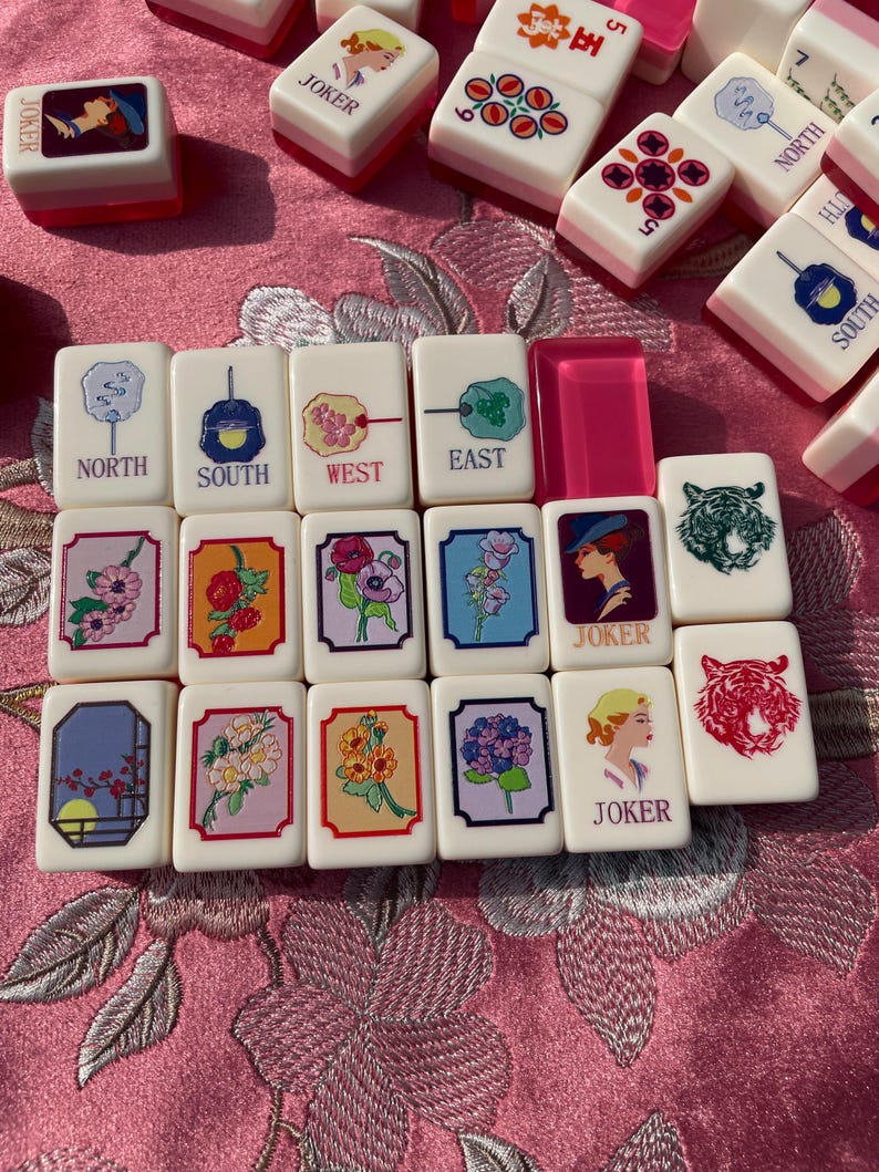 May include: A collection of vintage Mahjong tiles. The tiles are white with pink accents and feature various symbols, numbers, and characters. Some tiles display floral designs, while others have directional markers like "North" and "South". "Joker" tiles are also visible.