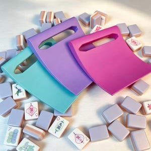 May include: Three curved, rectangular plastic pushers in teal, lavender, and magenta, each featuring a central rectangular cutout handle. These are scattered among a collection of small, rectangular mahjong tiles with white faces and lavender backs.