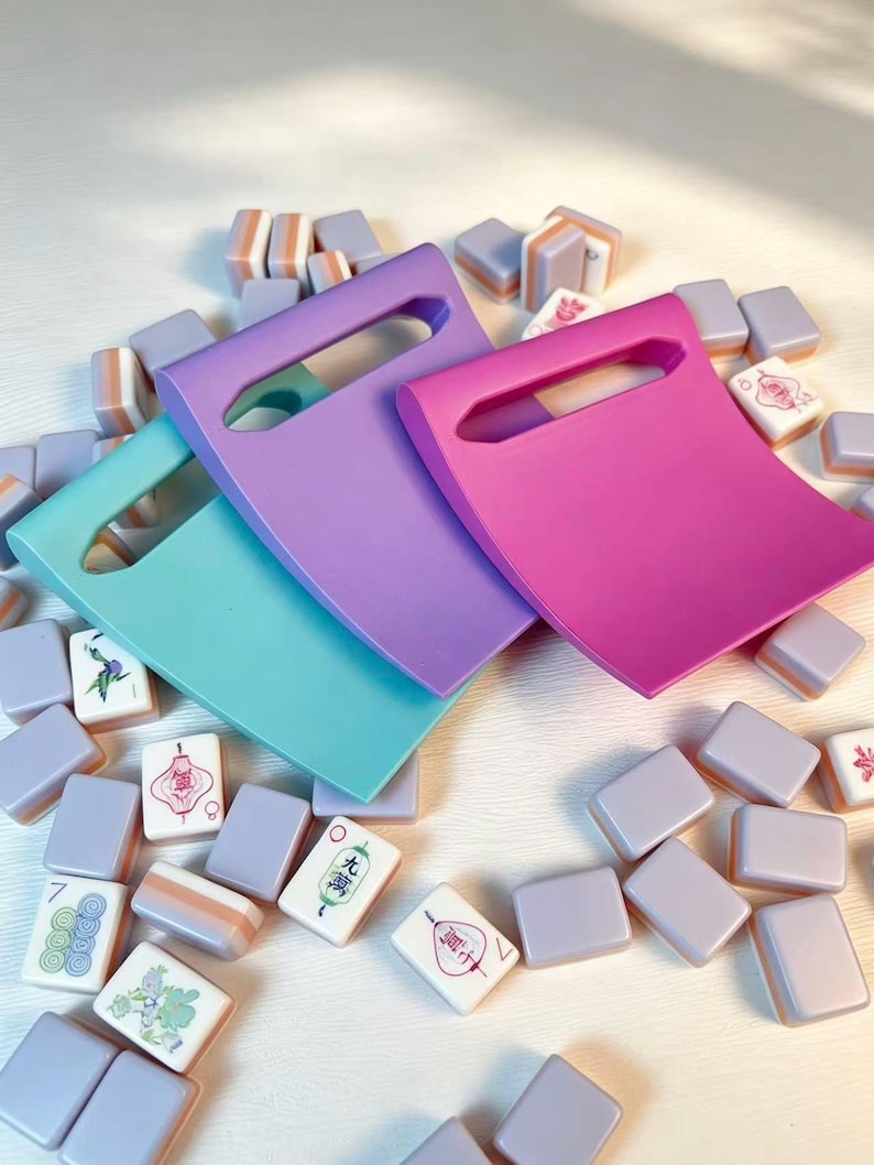 May include: Three curved, rectangular plastic pushers in teal, lavender, and magenta, each featuring a central rectangular cutout handle. These are scattered among a collection of small, rectangular mahjong tiles with white faces and lavender backs.