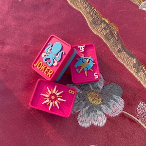 Puede incluir: Tres fichas de mahjong rectangulares de color rosa vibrante con bordes azules. Una ficha presenta un pulpo azul claro y el texto amarillo JOKER. Otra ficha muestra un s&iacute;mbolo azul y amarillo con el n&uacute;mero 5. La tercera ficha muestra un sol amarillo y el n&uacute;mero 8.