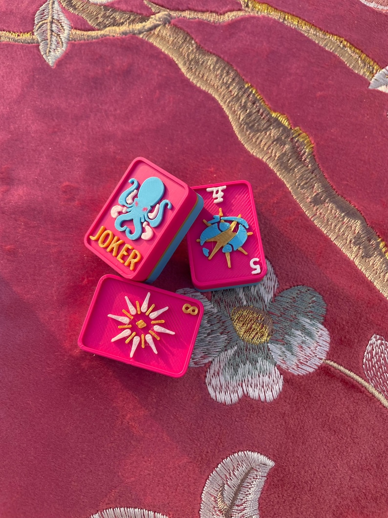 Puede incluir: Tres fichas de mahjong rectangulares de color rosa vibrante con bordes azules. Una ficha presenta un pulpo azul claro y el texto amarillo JOKER. Otra ficha muestra un s&iacute;mbolo azul y amarillo con el n&uacute;mero 5. La tercera ficha muestra un sol amarillo y el n&uacute;mero 8.