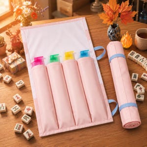 May include: A light pink fabric roll-up case featuring four vertical pockets, each containing a translucent plastic marker in pink, green, yellow, and blue. The case includes a matching light pink flap and a light blue strap for securing the roll.