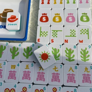 May include: A white Mahjong set with various tiles featuring symbols like horseshoes, money bags, and cacti. One tile reads "JOKER" with a cowboy hat. The set includes a blue tray.