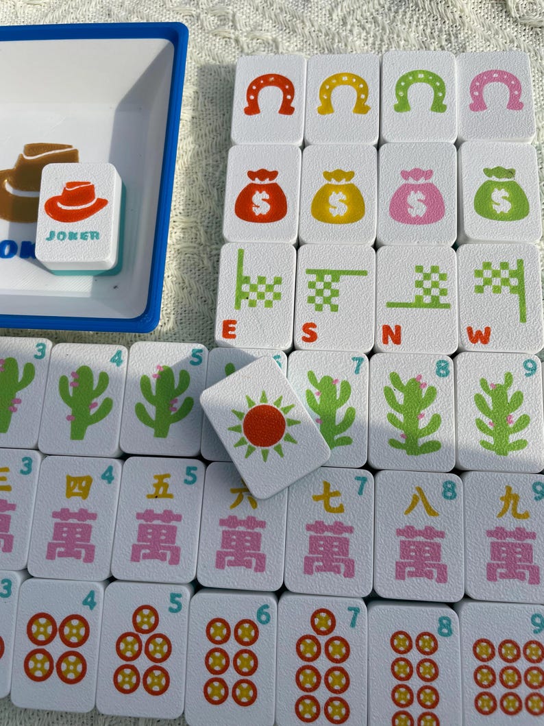 Może przedstawiać: Biały zestaw do gry w Mahjong z r&oacute;żnymi kafelkami przedstawiającymi symbole takie jak podkowy, worki z pieniędzmi i kaktusy. Jeden kafelek ma napis "JOKER" z kapeluszem kowbojskim. Zestaw zawiera niebieską tacę.
