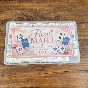 May include: A clear plastic storage case containing 160 white 3D printed American Mahjong tiles. The label on the lid features the text Floati MAHJ, Floating American Mahjong, 3D Printed, 160 Tiles, with illustrations of tiles showing a panda, bamboo, flowers, and a joker.