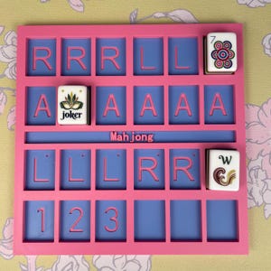 May include: A pink Mahjong tile holder with blue squares and pink letters. The word "Mah jong" is printed in pink. Several Mahjong tiles are in the holder, including a Joker tile and tiles with the numbers 7 and W.