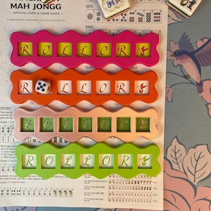 Pode incluir: Um colorido conjunto de jogos de Mah Jongg americano com quatro suportes de peças Rollor em rosa, laranja, rosa claro e verde. Cada suporte exibe a palavra "ROLLOR" e inclui um dado e peças de jogo. O guia do jogo é visível.