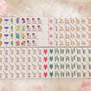 May include: A complete set of white Mahjong tiles, featuring various symbols, numbers, and characters. The tiles display illustrations of flowers, dragons, and jokers, with numbers from 1 to 9. The set is arranged on a soft, white surface.