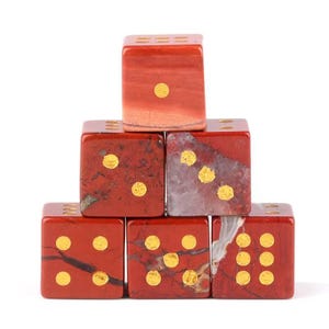 May include: A stack of six red jasper dice with gold-colored pips. The dice are arranged in a pyramid shape, with the top die showing one pip. The dice have a polished finish and a natural stone appearance.