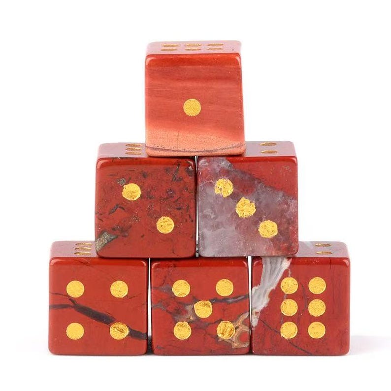 May include: A stack of six red jasper dice with gold-colored pips. The dice are arranged in a pyramid shape, with the top die showing one pip. The dice have a polished finish and a natural stone appearance.