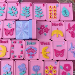 May include: Pink Mahjong tiles with various embossed designs in teal, yellow, and pink. The tiles feature floral, butterfly, and leaf motifs, along with numbers and directional symbols. A stack of matching tiles is also visible.