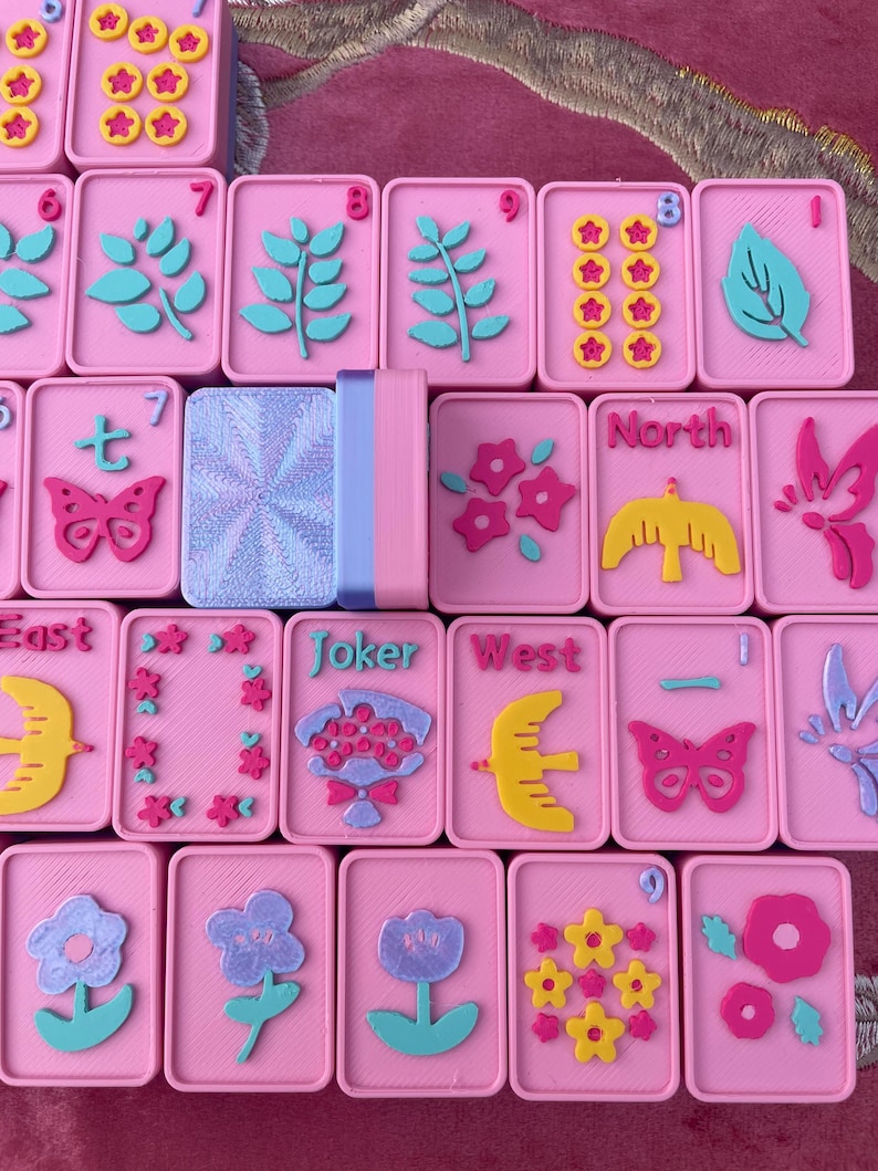 May include: Pink Mahjong tiles with various embossed designs in teal, yellow, and pink. The tiles feature floral, butterfly, and leaf motifs, along with numbers and directional symbols. A stack of matching tiles is also visible.