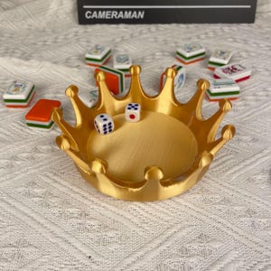 May include: A gold-colored crown-shaped tray with dice inside and Mahjong tiles scattered around. The tray has a circular base and pointed crown tips. The dice show the numbers 2 and 5. The text "CAMERAMAN" is visible in the background.