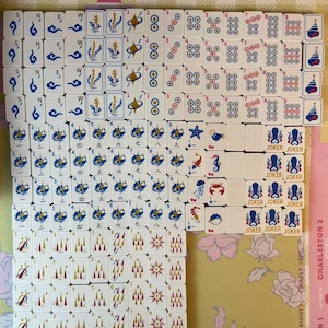 May include: A collection of white Mahjong tiles with blue, red, and gold symbols and numbers. The tiles are arranged on a floral patterned surface. Some tiles have the word "JOKER" printed on them.