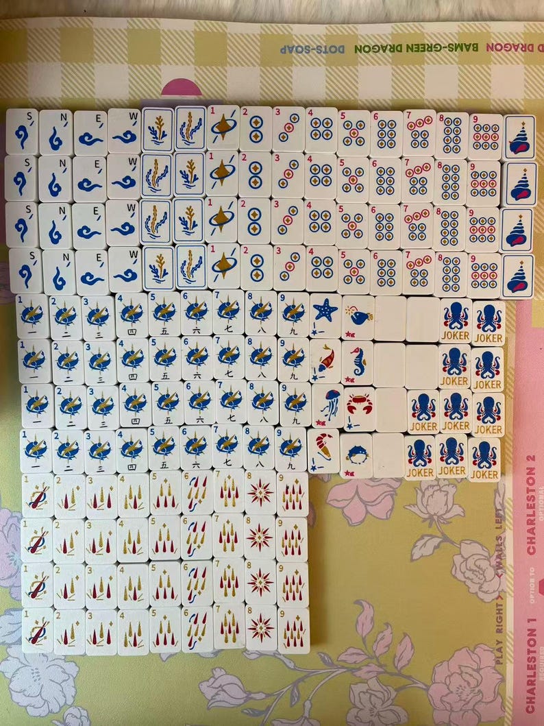 May include: A collection of white Mahjong tiles with blue, red, and gold symbols and numbers. The tiles are arranged on a floral patterned surface. Some tiles have the word "JOKER" printed on them.