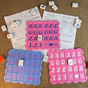 Op de afbeelding: Een afbeelding van drie kleurrijke Mahjongg-spellen in roze en blauw, elk met een geschulpte rand. De borden bevatten tegels met letters en cijfers, samen met spelinstructies en verspreide tegels. De spellen liggen op een geweven oppervlak.