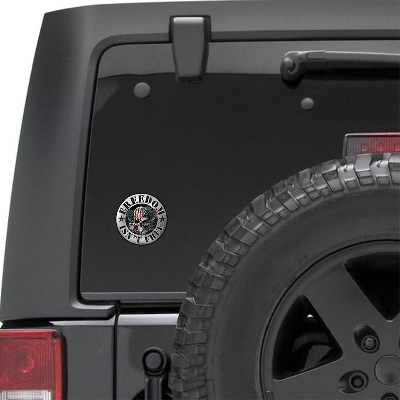 May include: A round sticker with the text "FREEDOM ISN'T FREE" encircling a skull design with an American flag motif. The sticker is affixed to the rear window of a black vehicle.