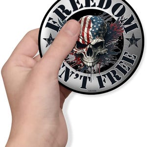 May include: A round sticker with a skull design. The skull is painted with the American flag and surrounded by the words "FREEDOM" and "AIN'T FREE". The sticker has a silver border and star accents.