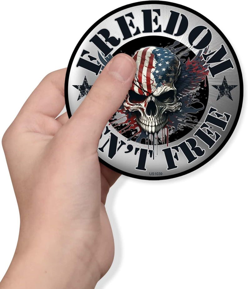 May include: A round sticker with a skull design. The skull is painted with the American flag and surrounded by the words "FREEDOM" and "AIN'T FREE". The sticker has a silver border and star accents.