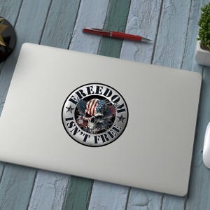 May include: A round sticker with the text "FREEDOM ISN'T FREE" encircling a skull with an American flag design. The skull has wings and is in the center of the sticker. The sticker is on a laptop.