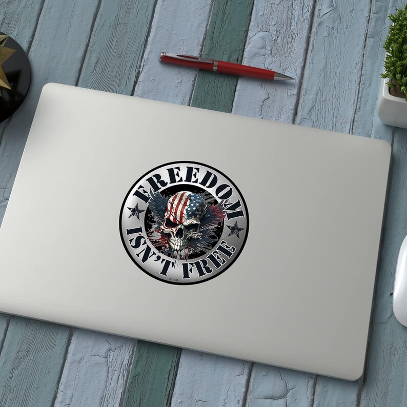 May include: A round sticker with the text "FREEDOM ISN'T FREE" encircling a skull with an American flag design. The skull has wings and is in the center of the sticker. The sticker is on a laptop.