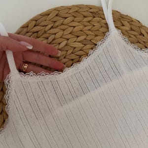 May include: White camisole with delicate lace trim along the neckline. The top features thin straps and a textured, vertically striped fabric with small diamond-shaped perforations. The camisole is displayed on a woven, circular surface.