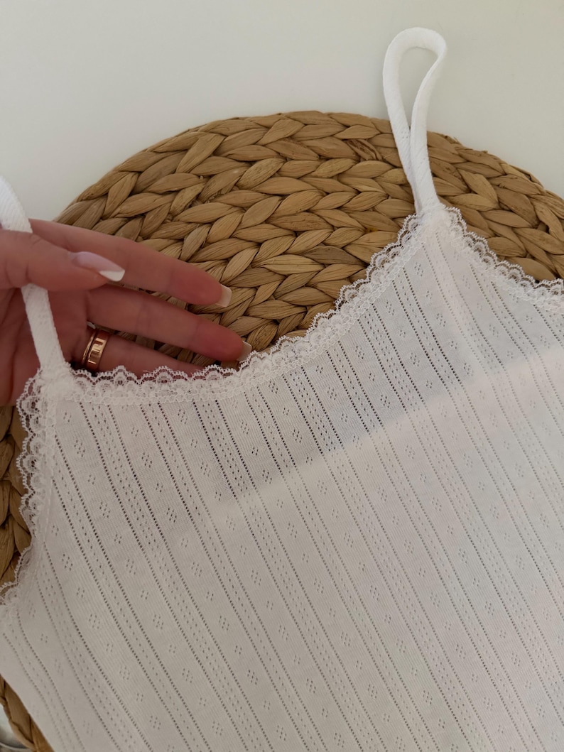 May include: White camisole with delicate lace trim along the neckline. The top features thin straps and a textured, vertically striped fabric with small diamond-shaped perforations. The camisole is displayed on a woven, circular surface.