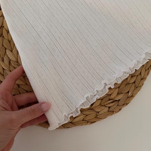 May include: Close-up of a white, textured fabric with vertical stripes and small diamond-shaped perforations. The edge of the fabric has a ruffled trim. The fabric is held by a hand, and rests on a woven, circular surface.