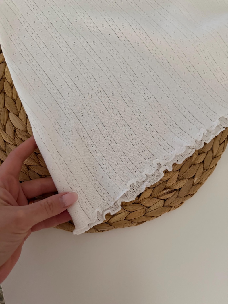 May include: Close-up of a white, textured fabric with vertical stripes and small diamond-shaped perforations. The edge of the fabric has a ruffled trim. The fabric is held by a hand, and rests on a woven, circular surface.