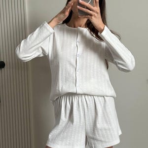 May include: A white ribbed knit pajama set. The set includes a long-sleeved button-down top and matching shorts. The fabric has a vertical ribbed texture. The shorts have an elastic waistband.