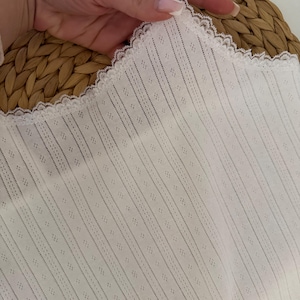 May include: White camisole with a delicate lace trim and thin straps. The fabric features a vertical striped pattern with small diamond-shaped perforations. The camisole is held up by a hand, showcasing the texture and design of the garment.