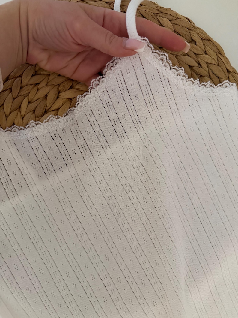 May include: White camisole with a delicate lace trim and thin straps. The fabric features a vertical striped pattern with small diamond-shaped perforations. The camisole is held up by a hand, showcasing the texture and design of the garment.