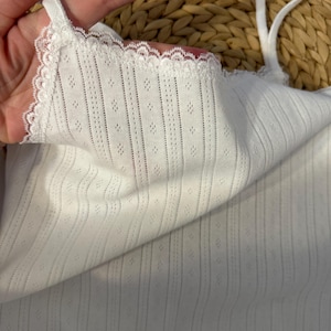May include: White camisole with delicate lace trim along the neckline and straps. The fabric has a textured, dotted pattern and vertical ribbed detailing. The camisole is a simple, elegant design.
