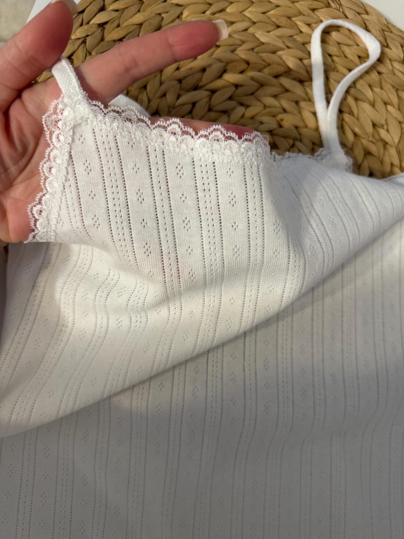 May include: White camisole with delicate lace trim along the neckline and straps. The fabric has a textured, dotted pattern and vertical ribbed detailing. The camisole is a simple, elegant design.