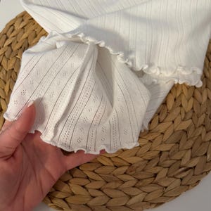 May include: Close-up of a white, textured fabric with a ruffled edge. The fabric has a delicate, openwork design with vertical stripes and small diamond-shaped perforations. The garment appears to be a top or sleeve.