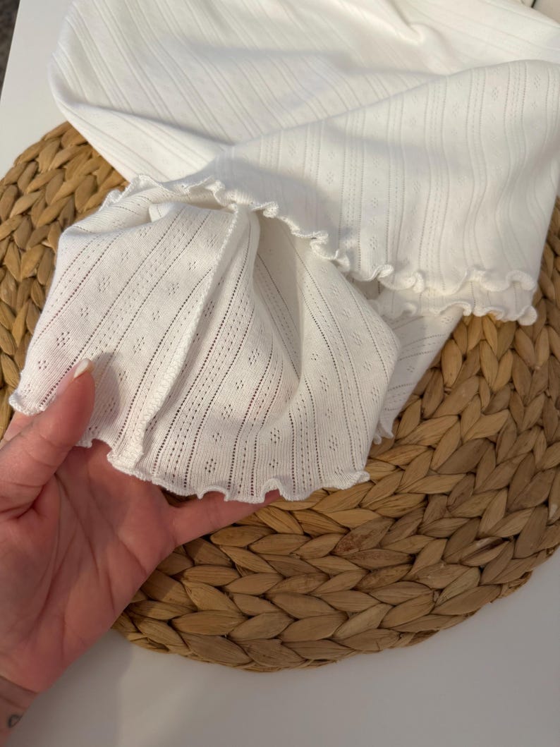 May include: Close-up of a white, textured fabric with a ruffled edge. The fabric has a delicate, openwork design with vertical stripes and small diamond-shaped perforations. The garment appears to be a top or sleeve.