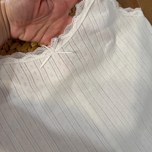 May include: White camisole with a delicate lace trim along the neckline and straps. The fabric features a subtle striped pattern with small diamond-shaped perforations. A small bow detail is centered at the neckline.