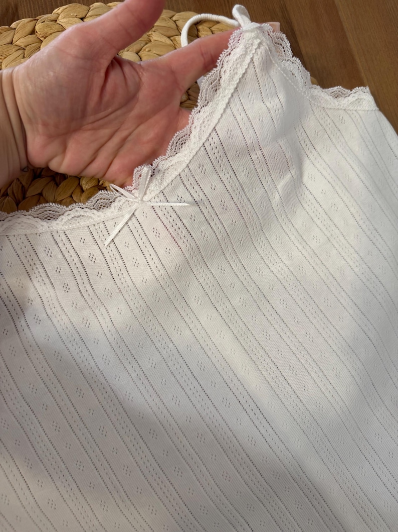 May include: White camisole with a delicate lace trim along the neckline and straps. The fabric features a subtle striped pattern with small diamond-shaped perforations. A small bow detail is centered at the neckline.