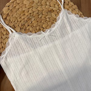 May include: A white camisole with thin straps and a delicate lace trim along the neckline. The fabric has a subtle ribbed texture with small diamond-shaped perforations. A small bow detail is centered on the front.