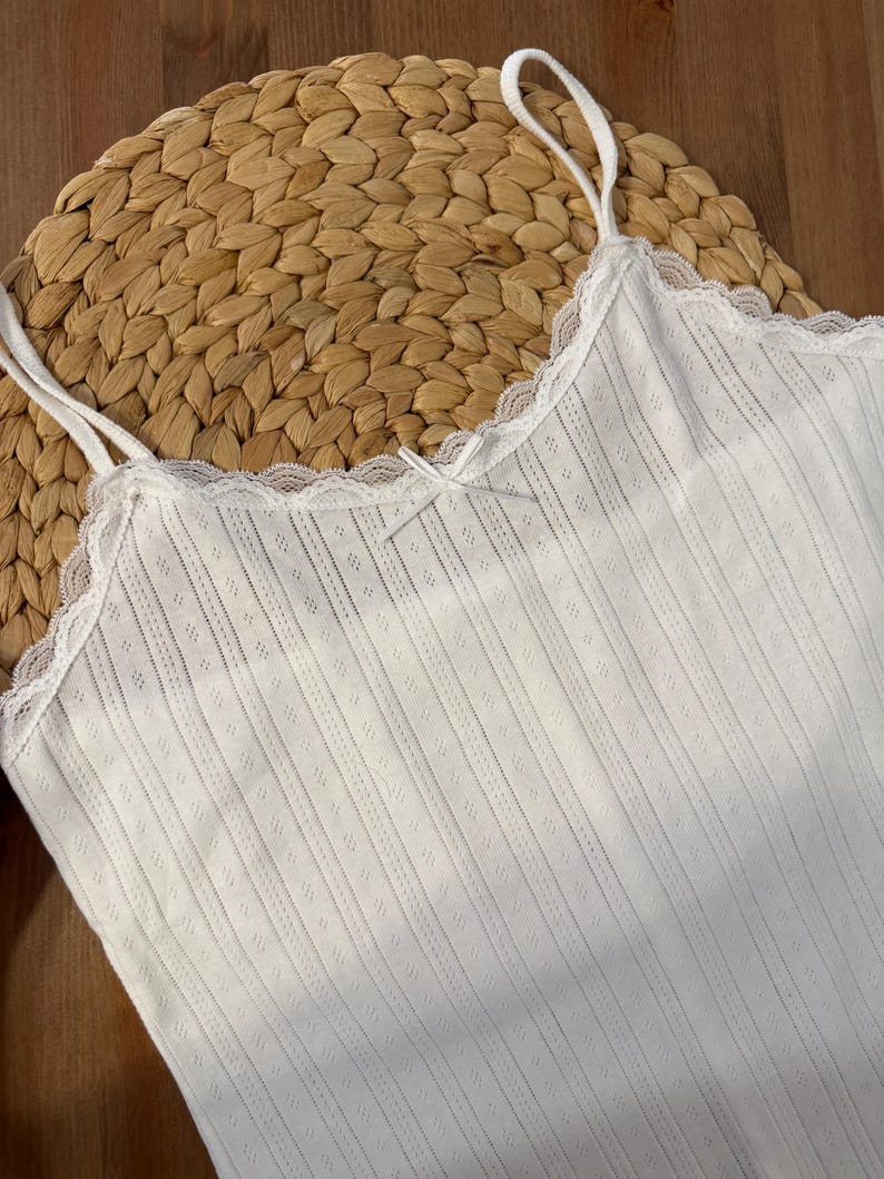 May include: A white camisole with thin straps and a delicate lace trim along the neckline. The fabric has a subtle ribbed texture with small diamond-shaped perforations. A small bow detail is centered on the front.