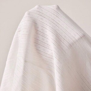 May include: Close-up of white fabric with a delicate, open weave. The fabric has a subtle texture with vertical and diagonal lines, creating a sense of depth and visual interest. The material appears soft and lightweight.